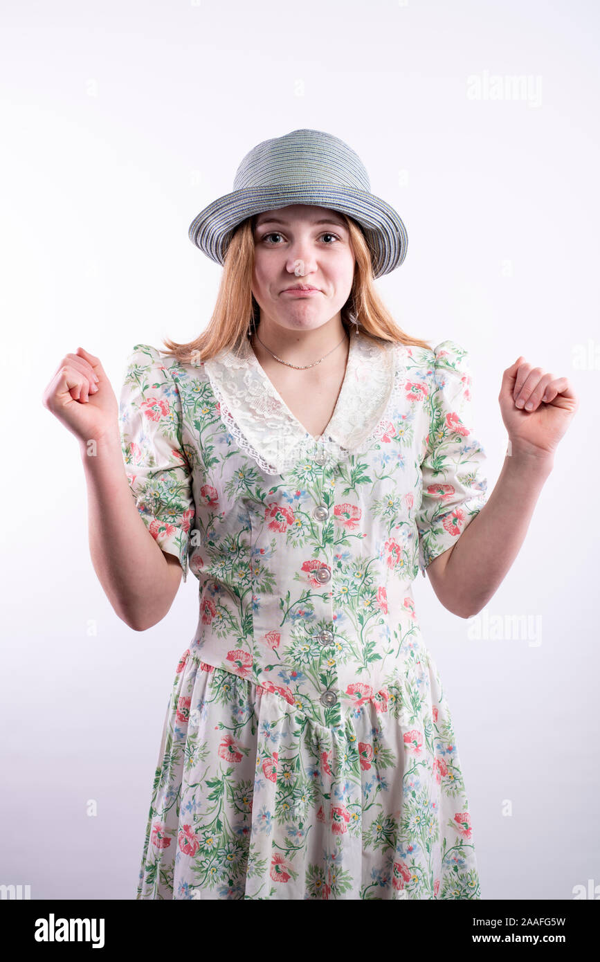 Caucasian teenage girl wearing a vintage floral dress and blue hat with an awkward expression against a white background, vertical orientation Stock Photo