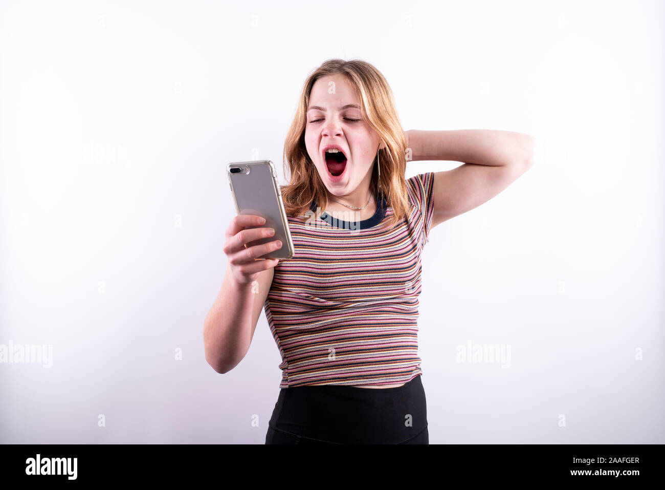 Caucasian teenage girl wearing a horizontal striped T-shirt and yawning with boredom while looking at a smartphone against a white background Stock Photo
