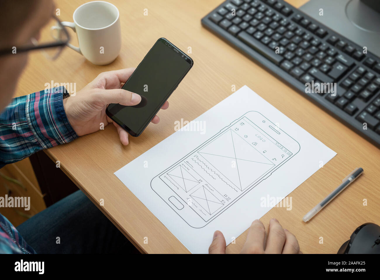 Designer sketches the layout of a responsive website or app and tests it on a mobile phone. Close up, work desk with computer beside. Stock Photo