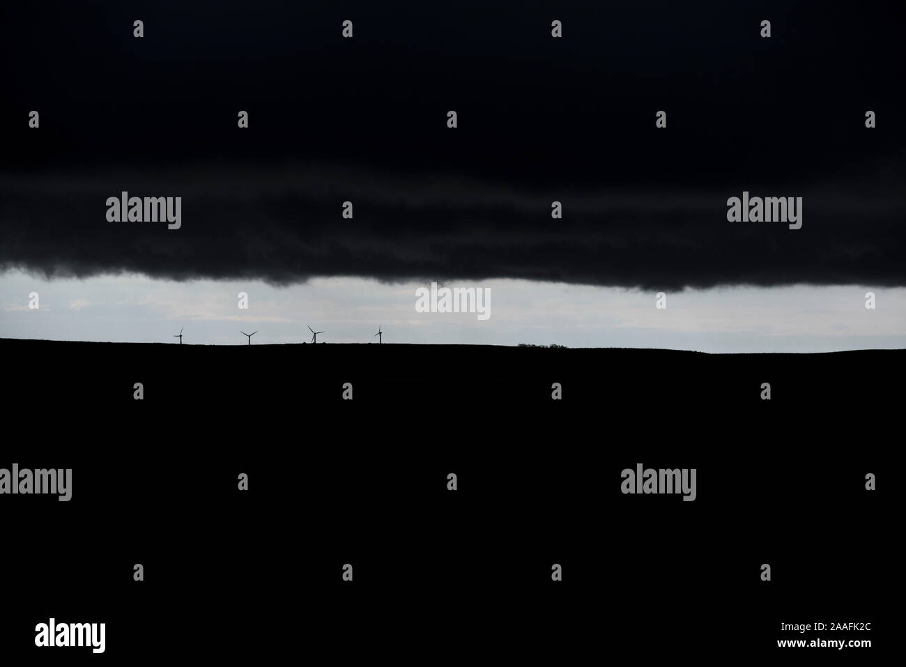 Distant wind turbines on the Canadian Prairies silhouetted against a thin band of blue sky with storm clouds overhead Stock Photo