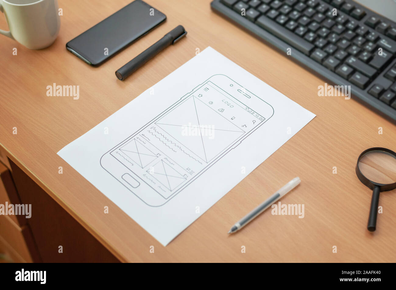 Sketch of a website or mobile phone app on wireframe. Paper on work desk. Keyobard, phone, pen, coffee and magnifier beside. Close up. Stock Photo