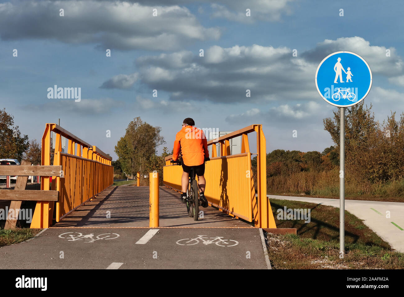 young man wearing striking orange jacket enjoying bicycle ride on early autumn sunny day, crossing steel orange cyclist / pedestrian bridge Stock Photo