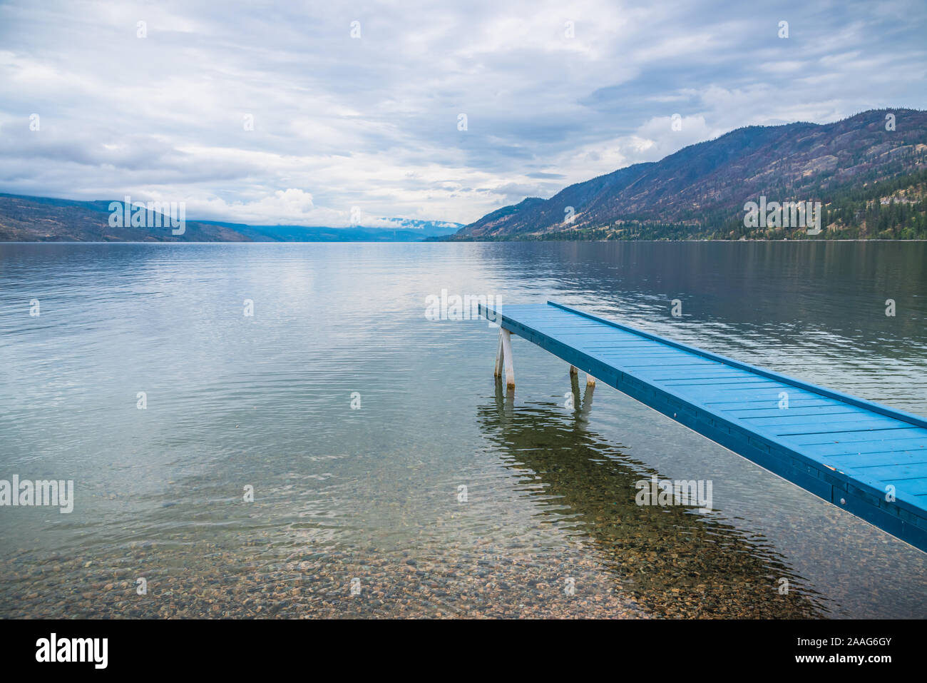 Vibrant blue painted dock extending over water toward view of mountains and overcast sky at Okanagan Lake Stock Photo