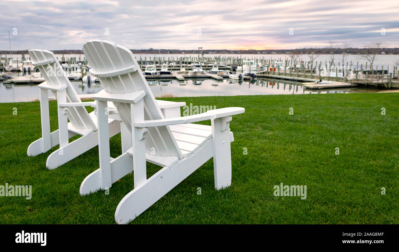 White "Adirondack" style lounge chairs on green grass at waterfront marina, with boats docked in background Stock Photo