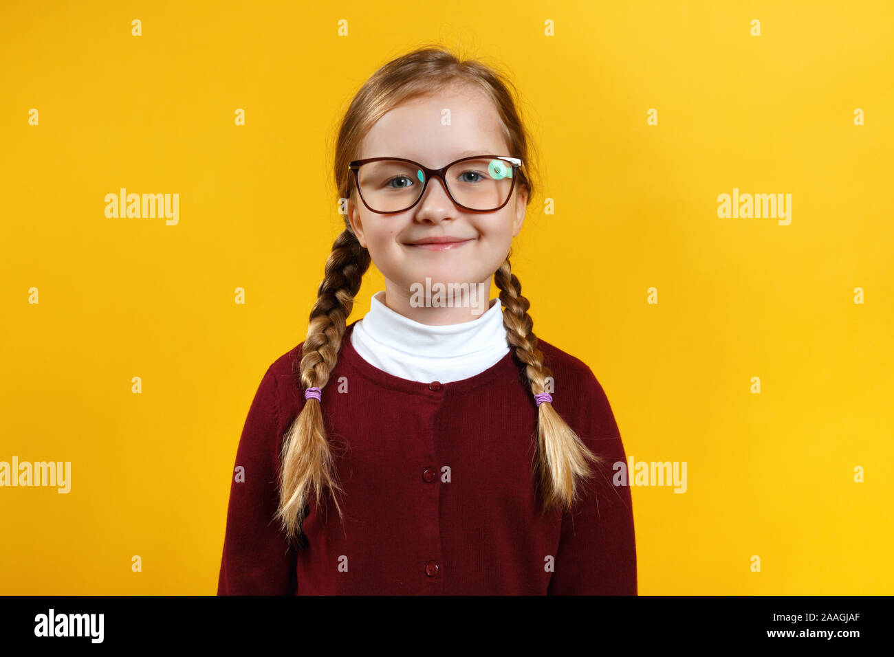 Smart girl schoolgirl with glasses and pigtails. A child in a red sweater on a yellow background. Stock Photo