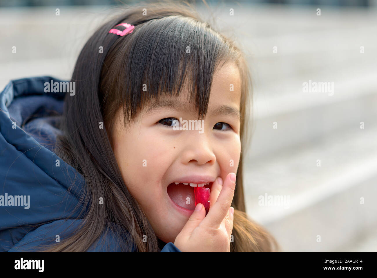 Pretty little Asian girl popping a colorful red sweet into her mouth while glancing sideways at the camera Stock Photo