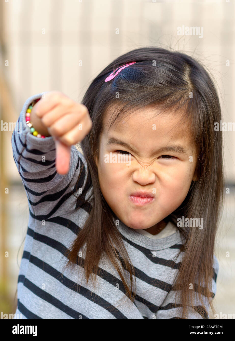 Portrait of a angry little Asian girl giving a thumb down gesture at the camera while pulling a fierce face in a show of childish aggression Stock Photo