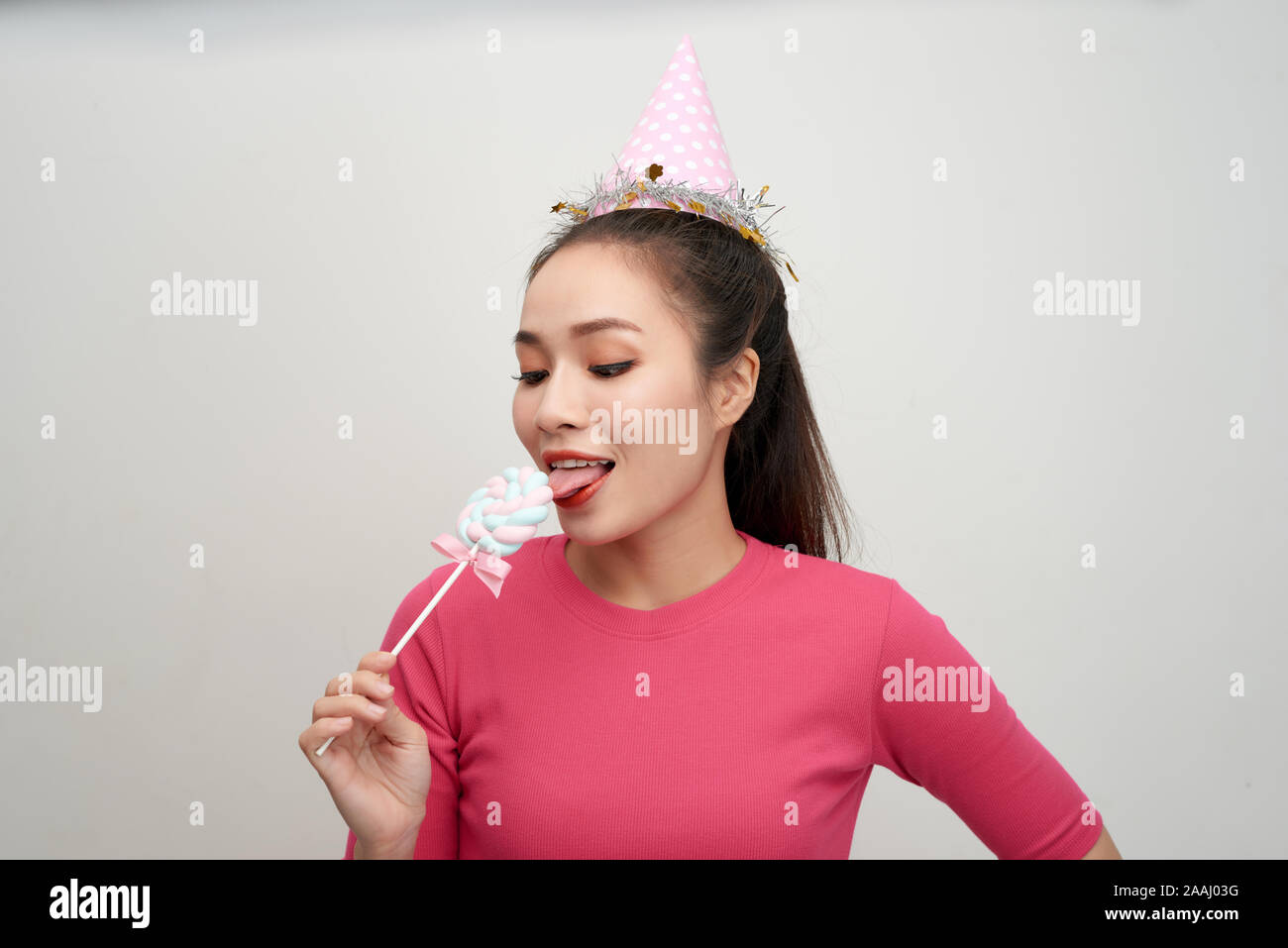 Blinking young woman in birthday hat holding, licking round lollipop celebrating Stock Photo