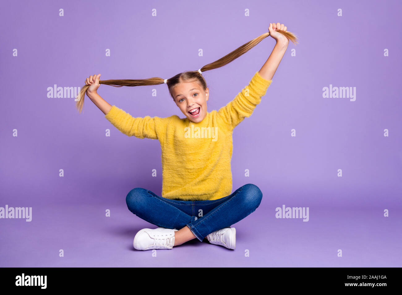 Full length photo of cheerful content child sit crossed folded legs have fun on weekends hold her ponytails wear casual style outfit isolated over Stock Photo