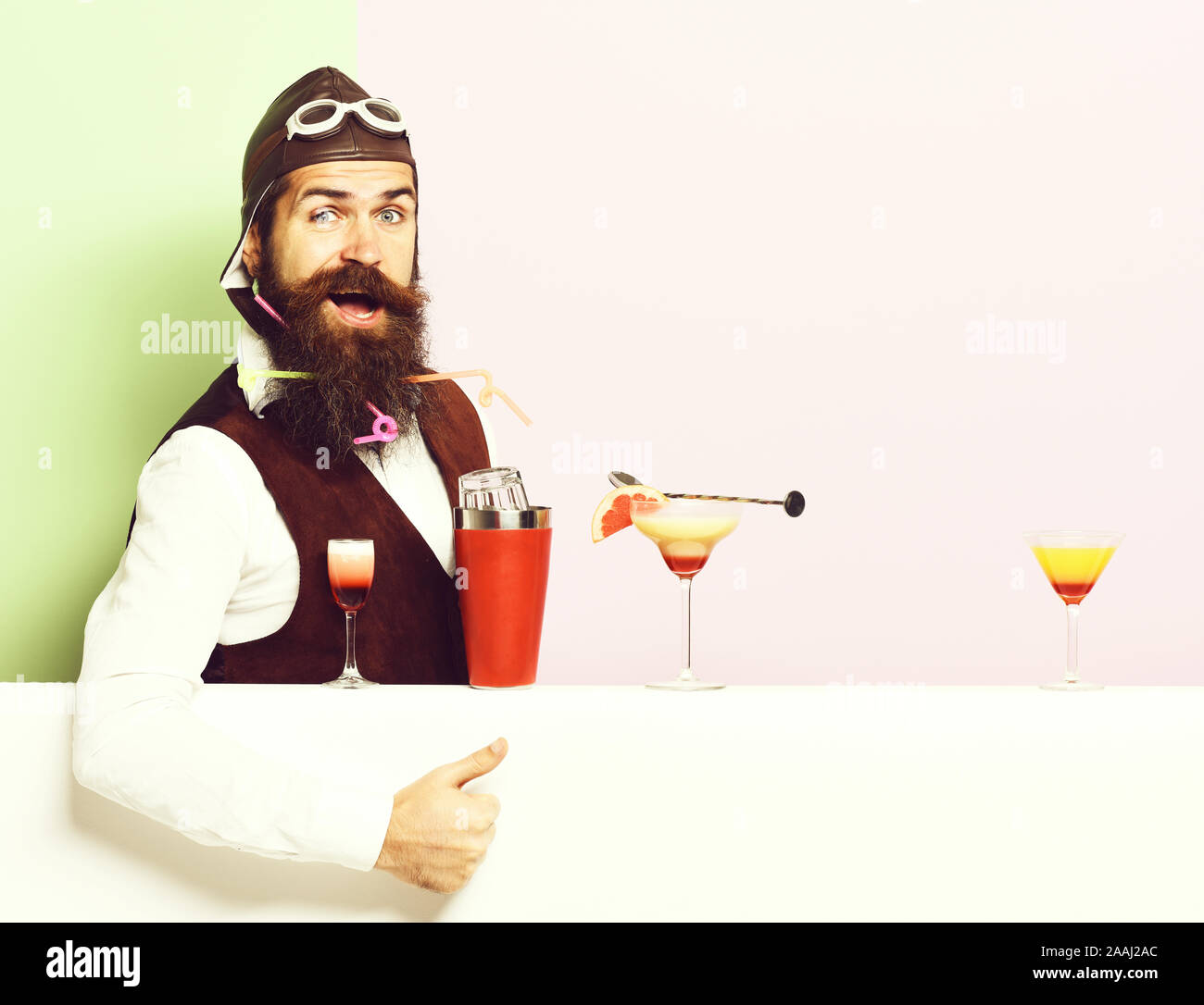 handsome bearded aviator or pilot with beard and mustache on smiling face with shaker alcoholic cocktails in vintage suede leather waistcoat with hand and glasses. on purple green studio background Stock Photo