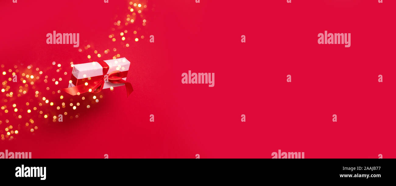 Big gift box wrapped in wrapping pink paper red satin ribbon bow, with shiny glitter flying in the air on a red background with copy space. Stock Photo