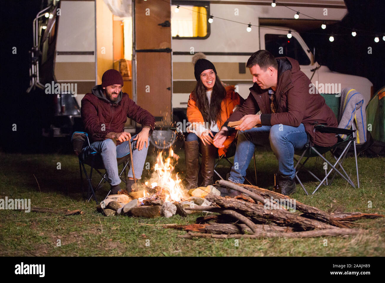 Group of friend relaxing and laughing together around camp fire in the mountains. Retro camper van. Stock Photo