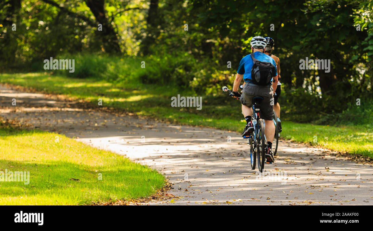 Actively aging, two senior bicyclists, a man and woman, riding away from the camera, framed right, in bright dappled sunlight; copy space left. Stock Photo