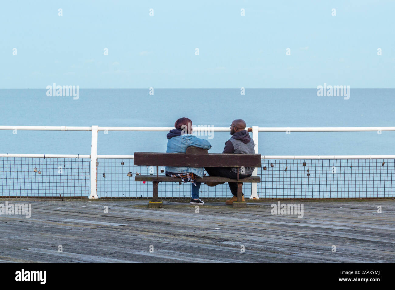 A man and woman sit talking on a bench while looking out to sea. Love locks are attached to the railing in front of them. Stock Photo