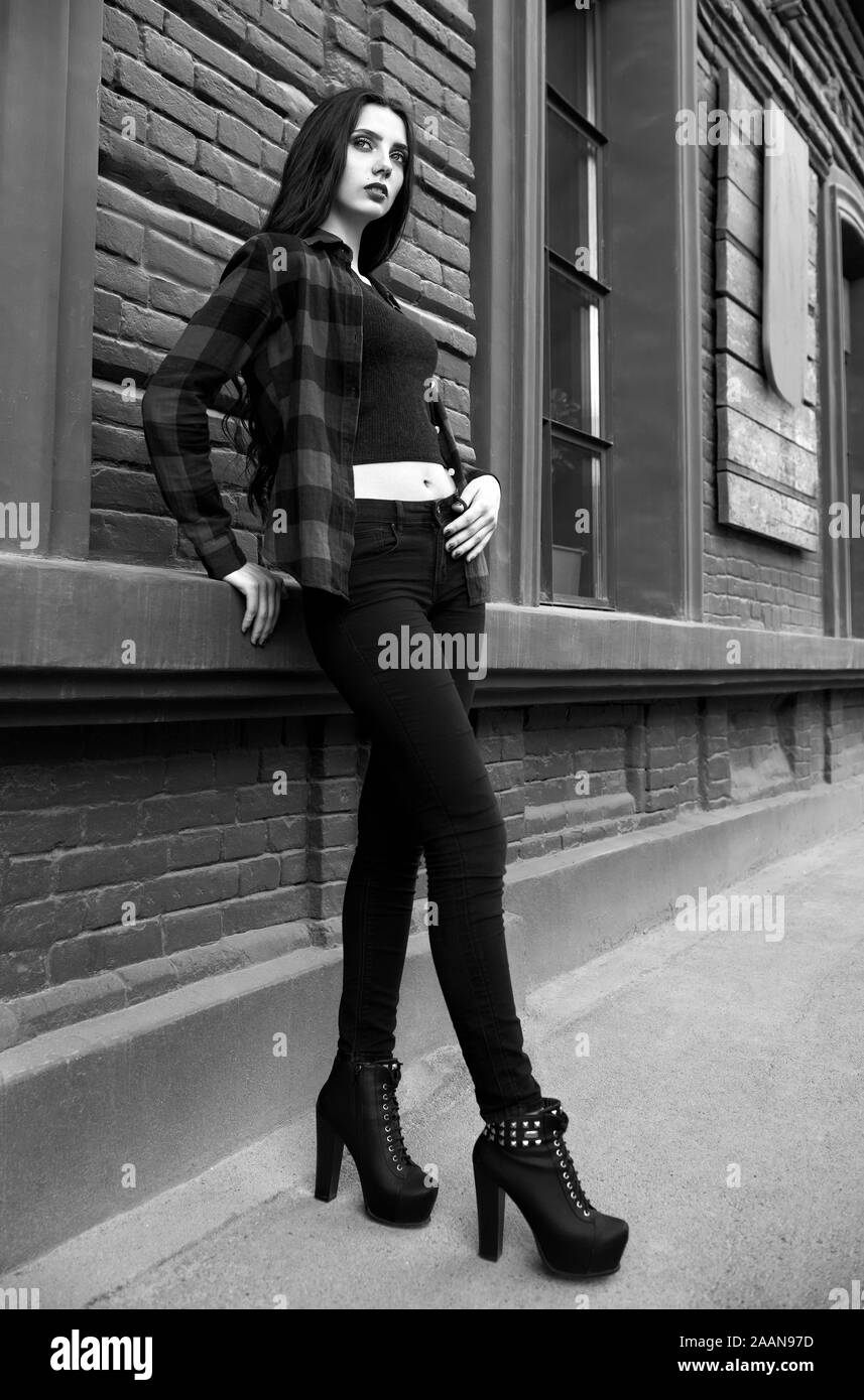 Street fashion: beautiful young girl wearing checkered shirt, jeans, topic and shoes stands at the wall. Black and white Stock Photo