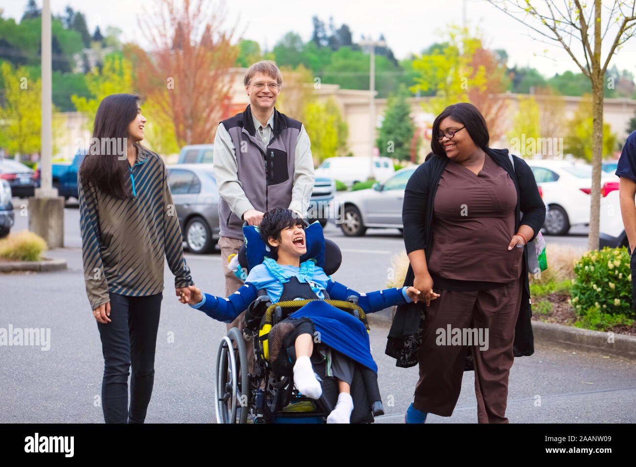 Disabled biracial boy in wheelchair walking along city street with father, siblings and caregiver, holding hands Stock Photo