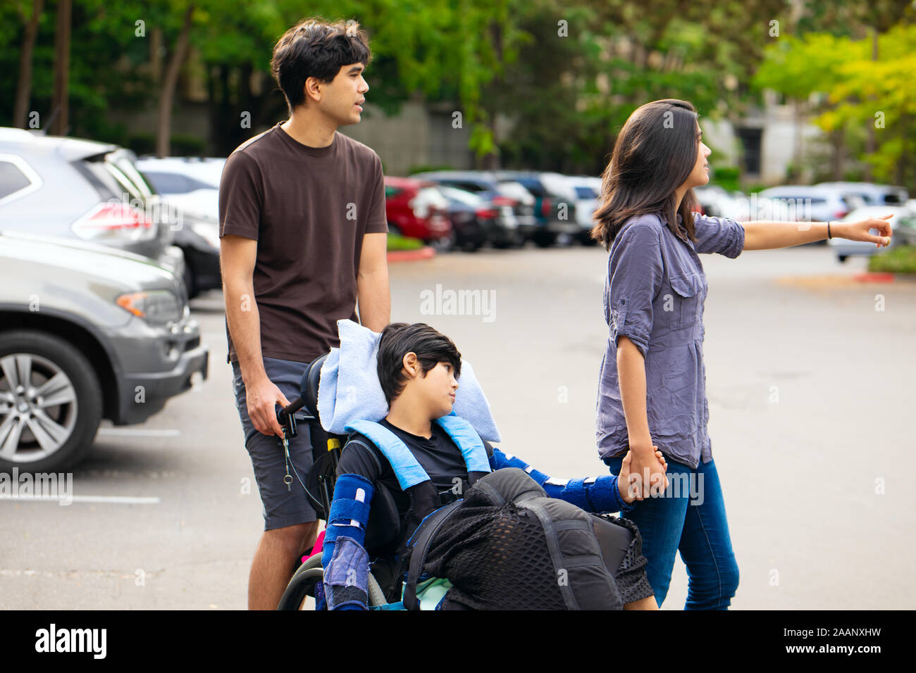 Older brother and sister pushing disabled little boy in wheelchair across parking lot Stock Photo