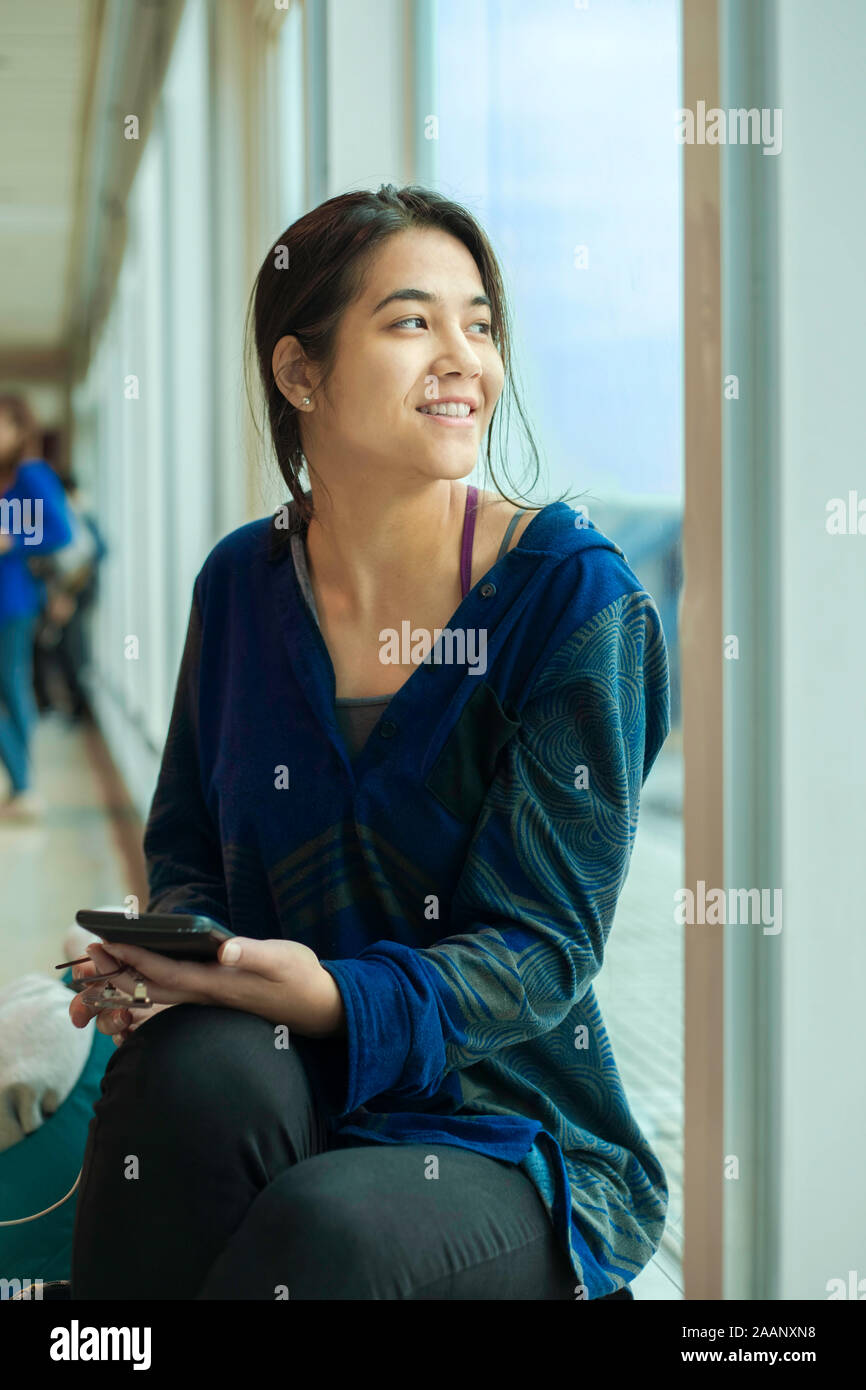 Teen girl or young woman sitting along large sunny windows at airport waiting for flight, looking out window and holding cell phone Stock Photo