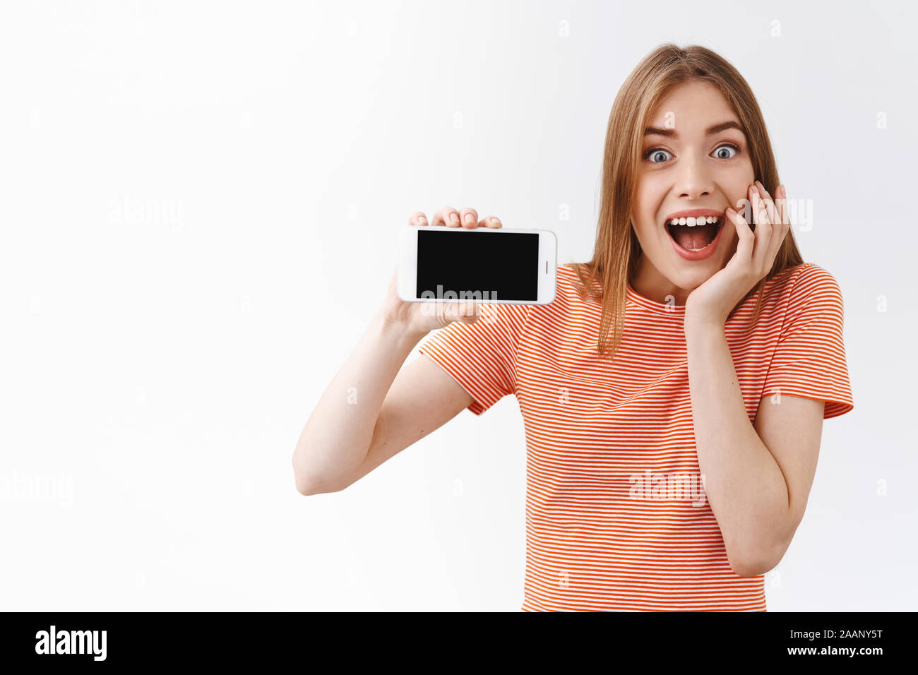 Surprised, eager and enthusiastic good-looking young caucasian woman in striped t-shirt, holding smartphone horizontally, showing black mobile display Stock Photo