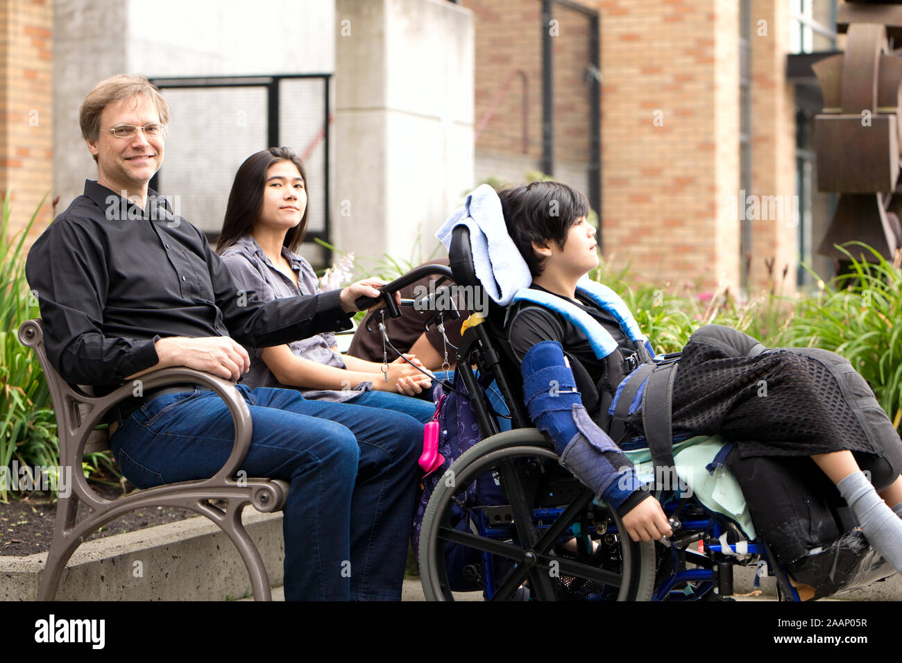 Multiracial family with special needs child sitting outdoors together on summer day. Child is sitting in wheelchair. Stock Photo