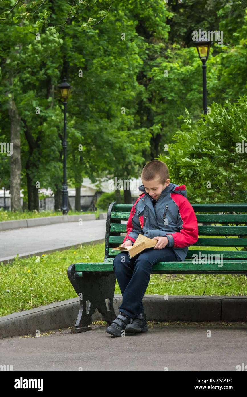 Smiling boy in casual clothing is reading a book. Boy is sitting alone on old wooden bench in park. Selective focus. Unfocused green park at backgroun Stock Photo