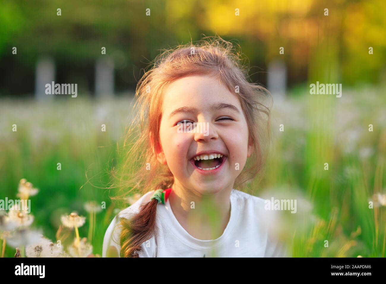 Beautiful little girl  laughing and playing with flowers in sunny spring park. Happy cute kid having fun outdoors at sunset. Stock Photo