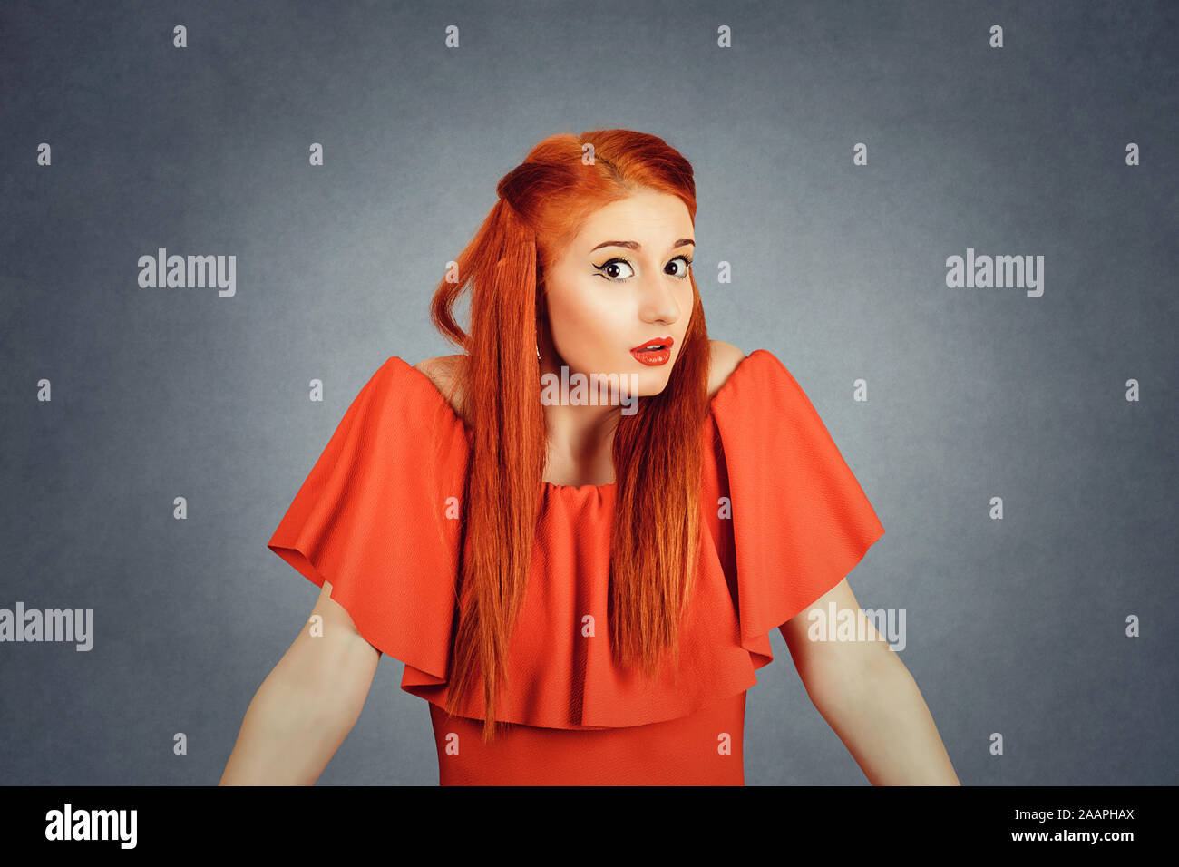 Confused young redhead woman looking at you camera, shrugging shoulders. Young redhead lady wearing red dress and yellow makeup isolated on Red backgr Stock Photo