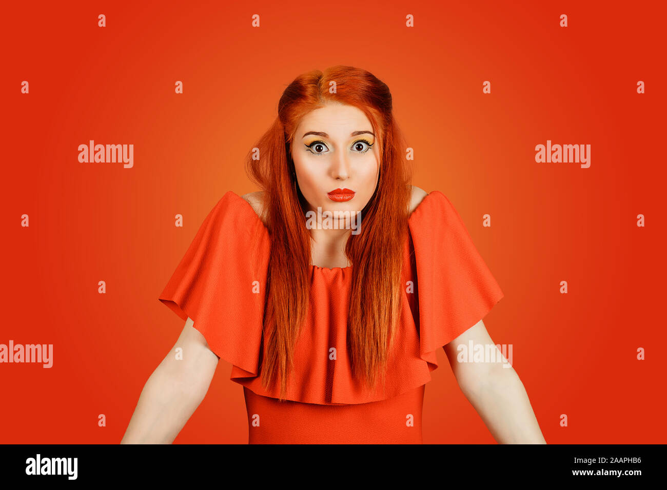 Confused guilty looking woman shrugging shoulders in confusion. Young redhead lady wearing red dress and yellow makeup isolated on Red background Stock Photo