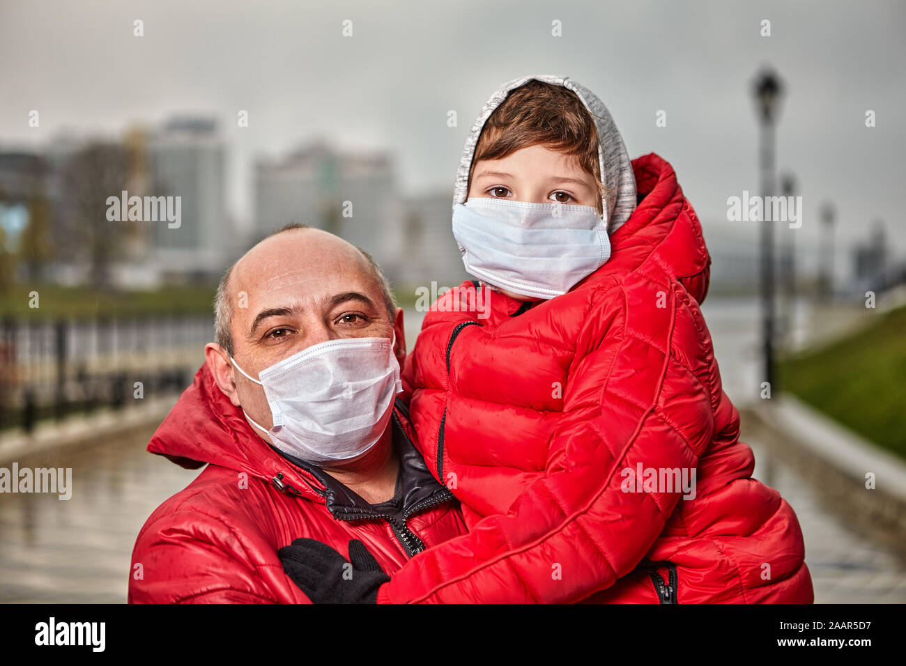 A middle-aged man, over 50 years old, holds a 7-year-old baby boy in his arms, both are dressed in warm clothes and cover their faces with protective Stock Photo