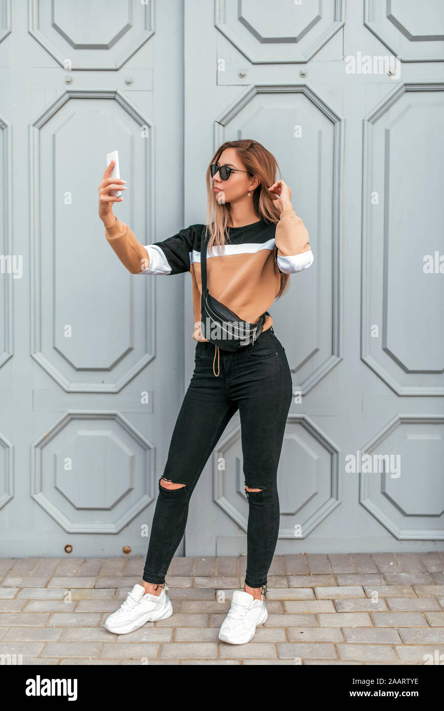 Beautiful fashionable and stylish woman summer city. Photographs himself on  phone, selfie on camera, modern hipster clothes, sweaters and jeans Stock  Photo - Alamy