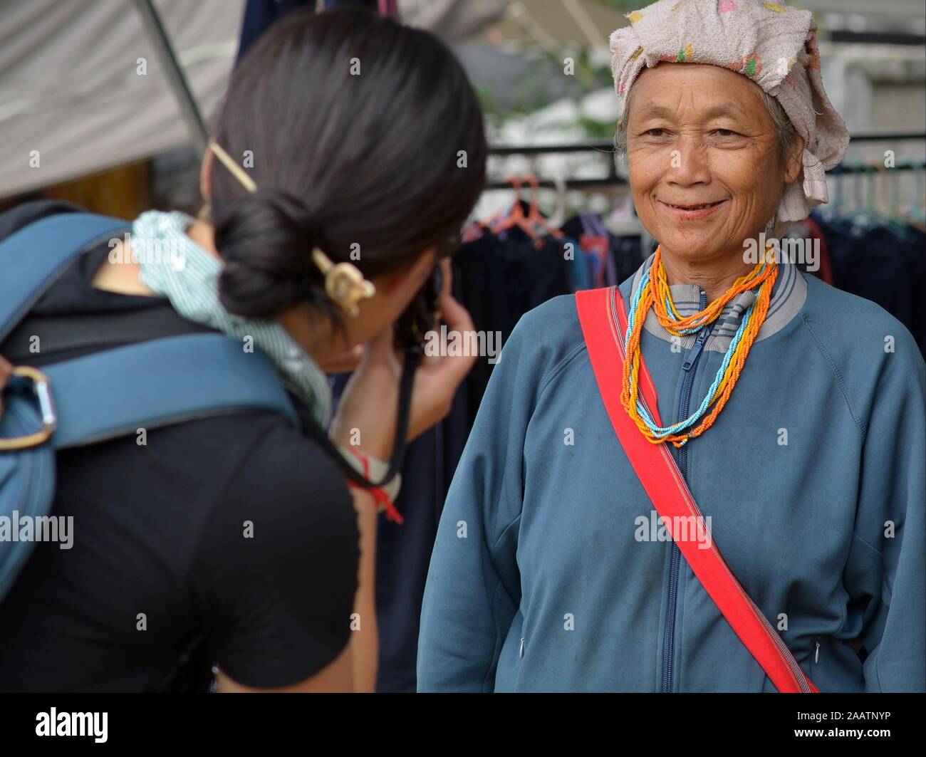 Japanese portrait photographer (tourist) takes a portrait photo of an elderly Thai Karen tribal woman with betel-nut stained lips at weekly market. Stock Photo