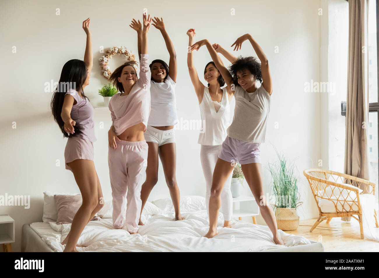 Happy multiethnic women wear pyjamas dancing on bed together Stock