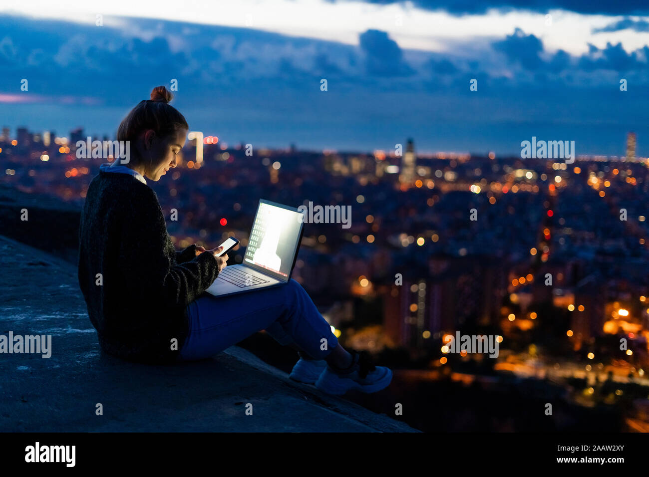 Young woman using cell phone and laptop at dawn above the city, Barcelona, Spain Stock Photo