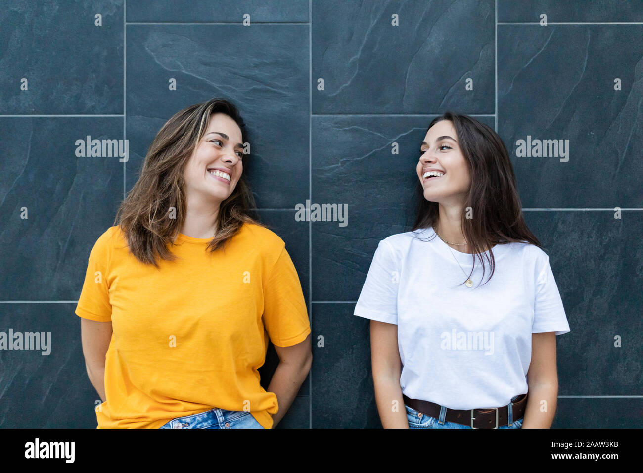 Two friends leaning on a gray wall and laughing Stock Photo