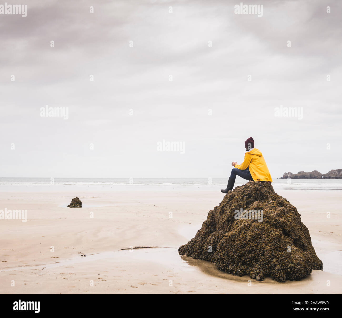Young woman wearing yellow rain jacket sitting on rock at the beach ...