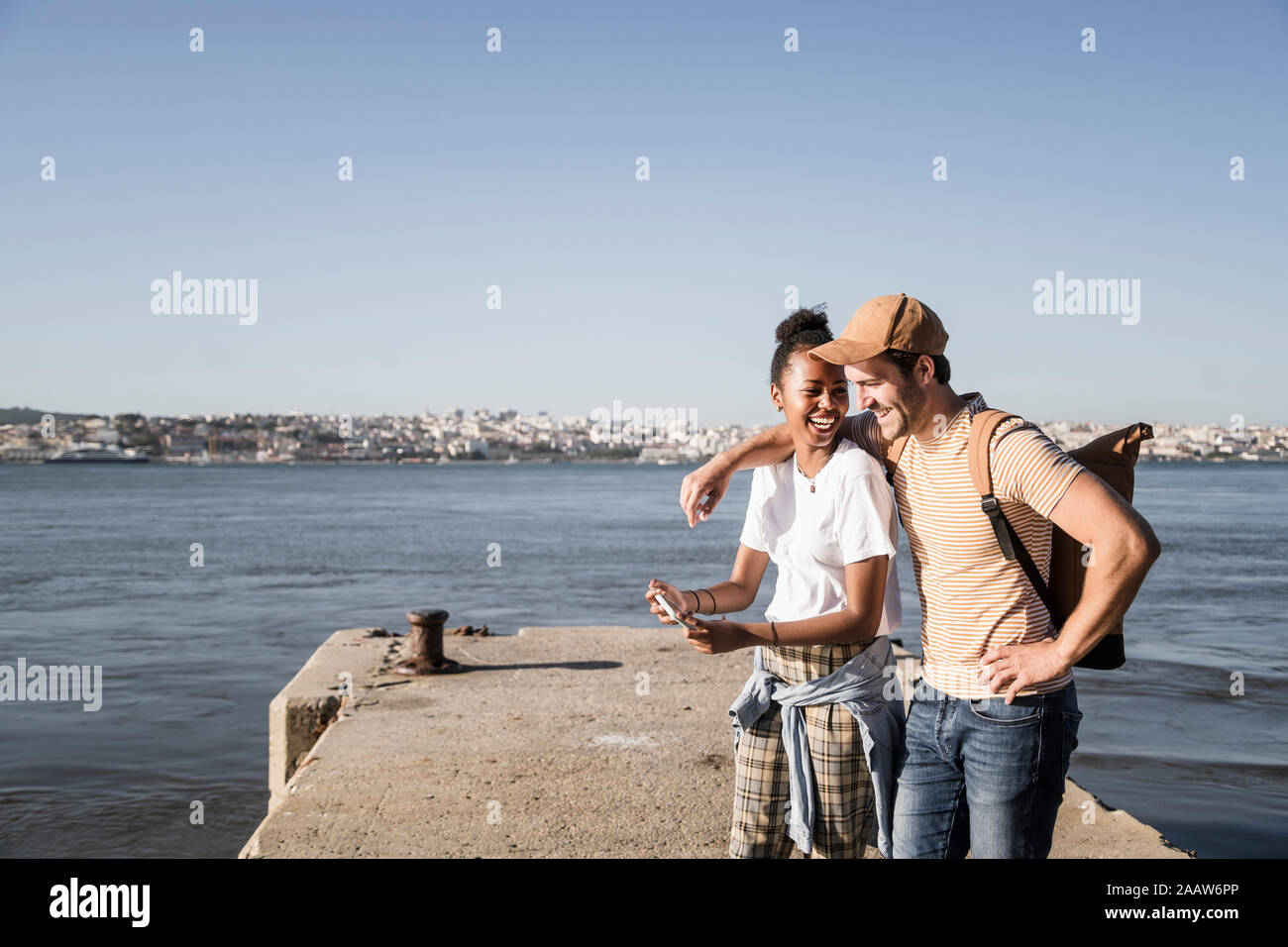 Happy young couple using cell phone on pier at the waterfront, Lisbon, Portugal Stock Photo