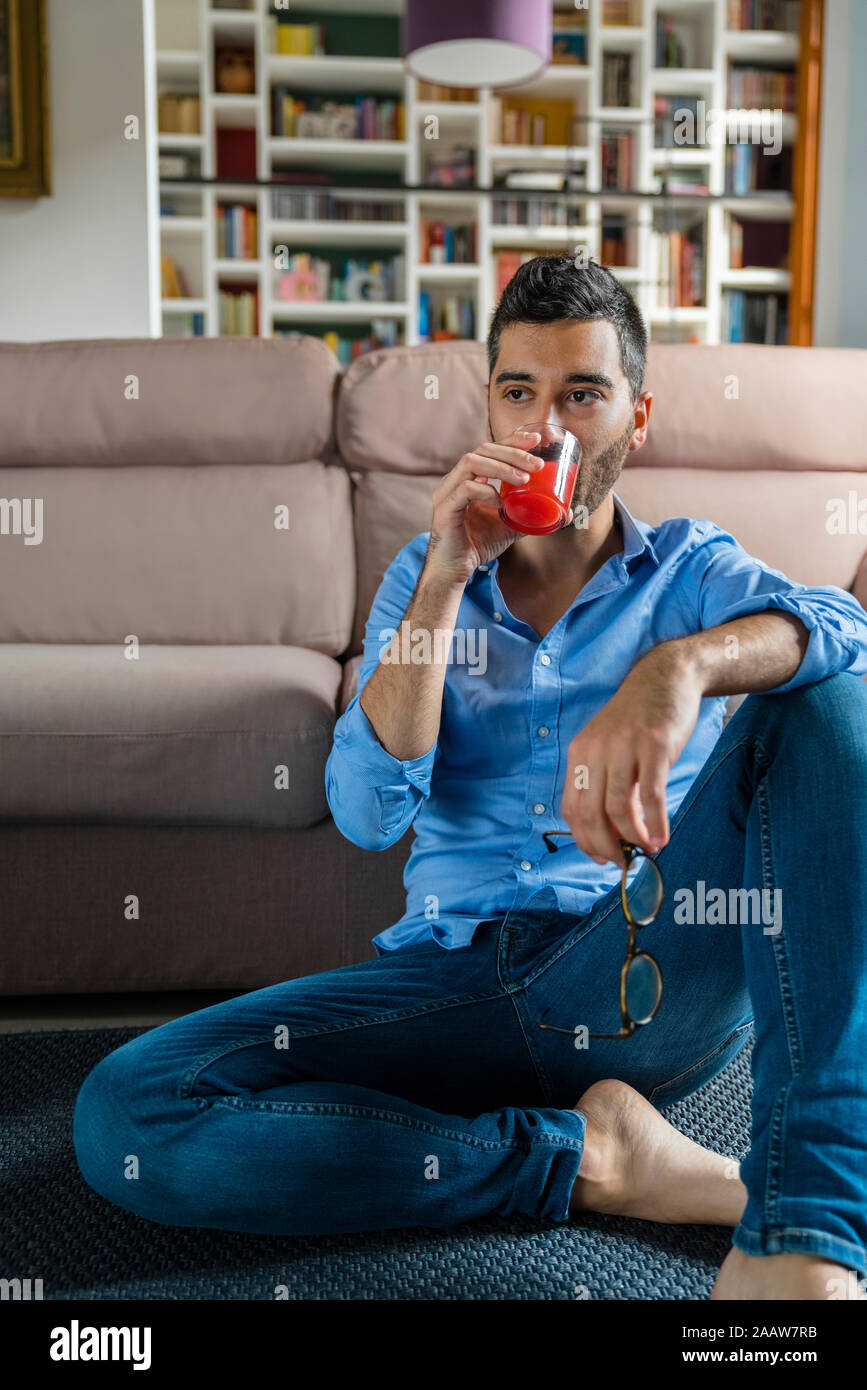 Portrait of young man sitting on the floor at home drinking glass of juice Stock Photo