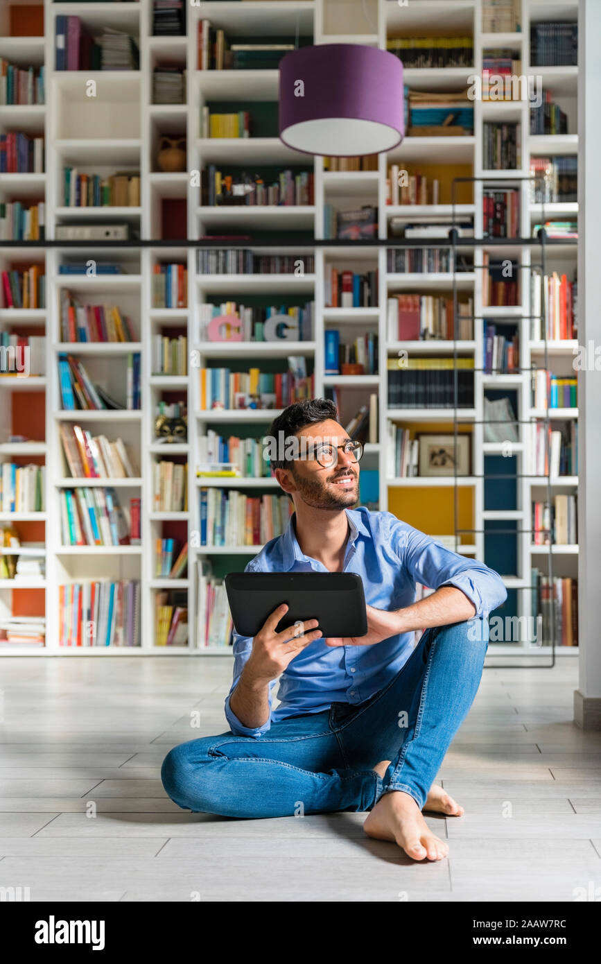 Portrait of barefoot young man sitting in front of bookshelves on the floor with digital tablet Stock Photo