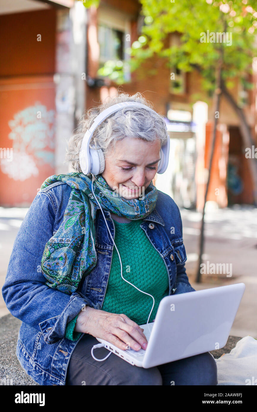 Senior woman with headphones using laptop in the city Stock Photo