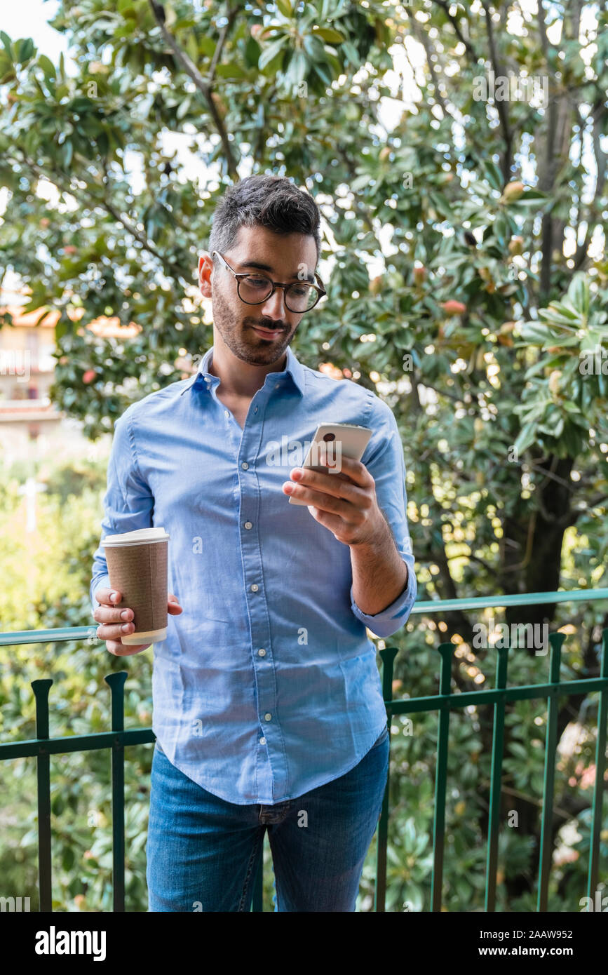 Portrait of young man standing on balcony with coffee to go looking at cell phone Stock Photo
