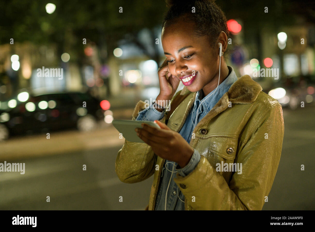 Portrait of smiling young woman using earphones and smartphone in the city by night, Lisbon, Portugal Stock Photo