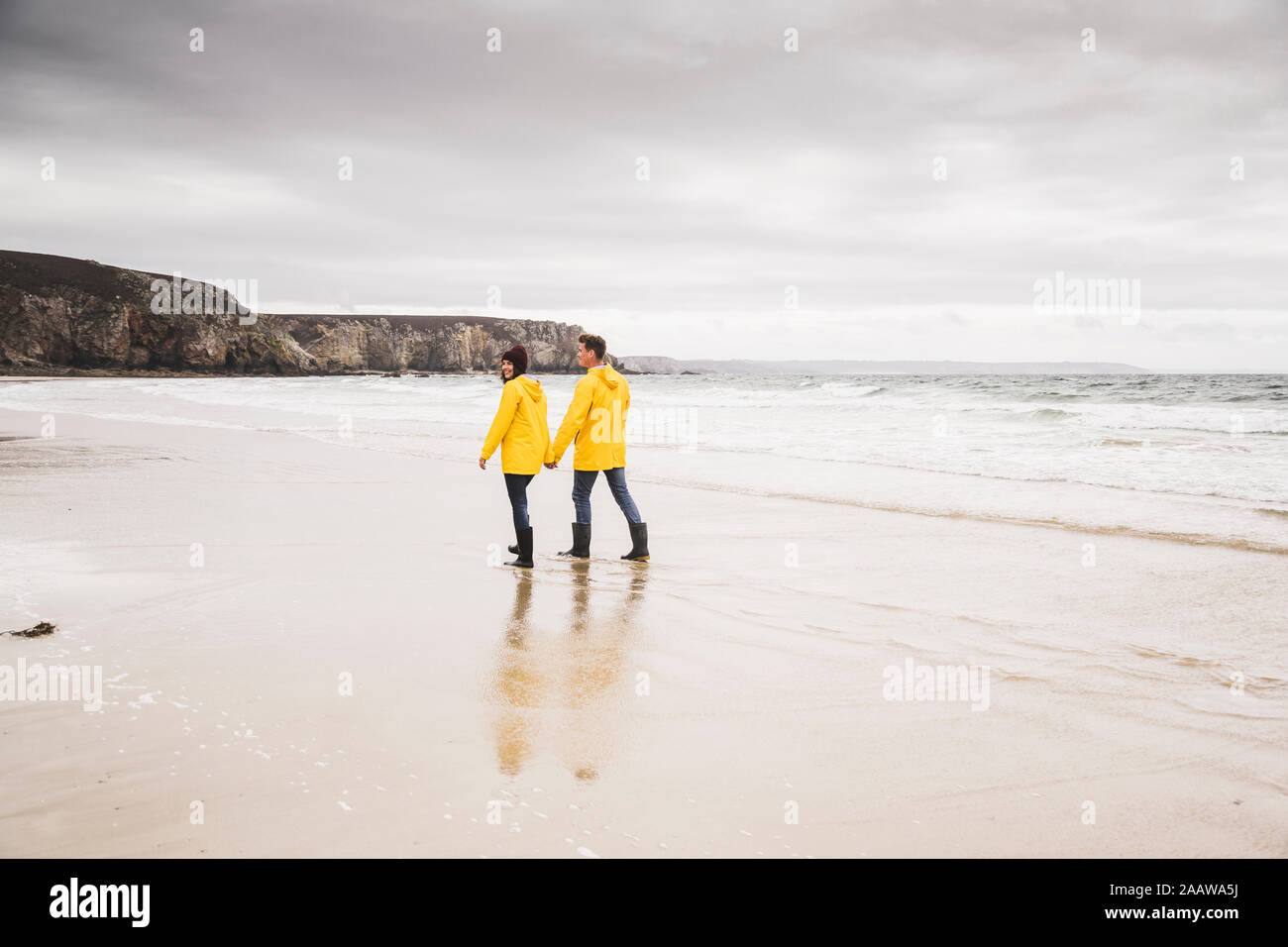 Young woman wearing yellow rain jackets and walking along the beach, Bretagne, France Stock Photo