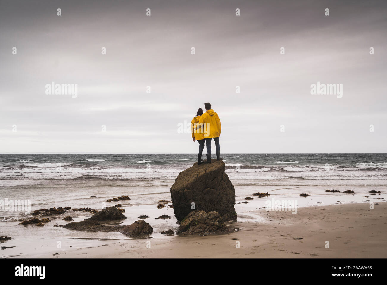 Young woman wearing yellow rain jackets and standing on rock at the beach, Bretagne, France Stock Photo