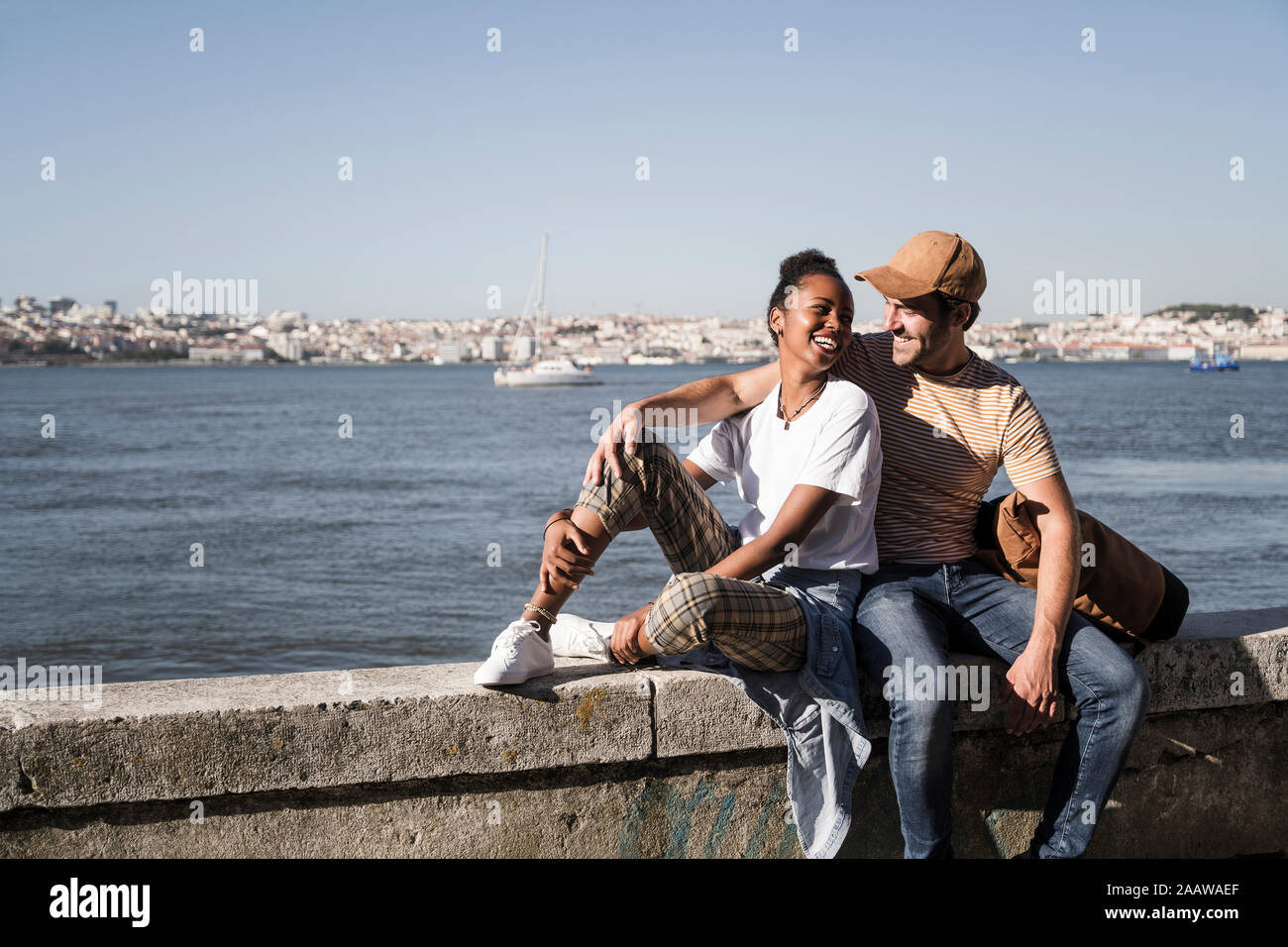 Happy young couple sitting on a wall at the waterfront, Lisbon, Portugal Stock Photo
