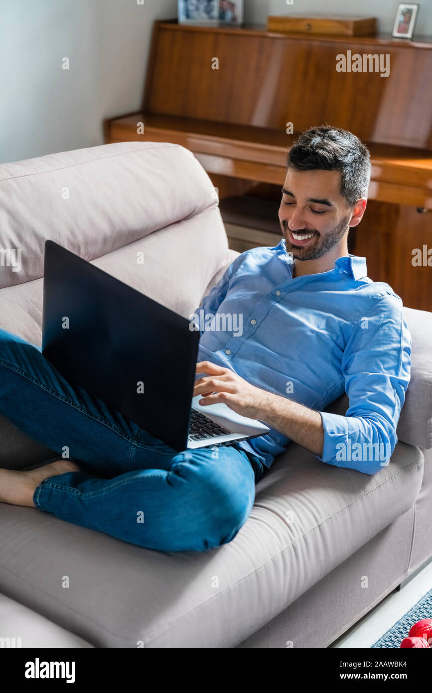 Smiling young man sitting on the couch at home using laptop Stock Photo
