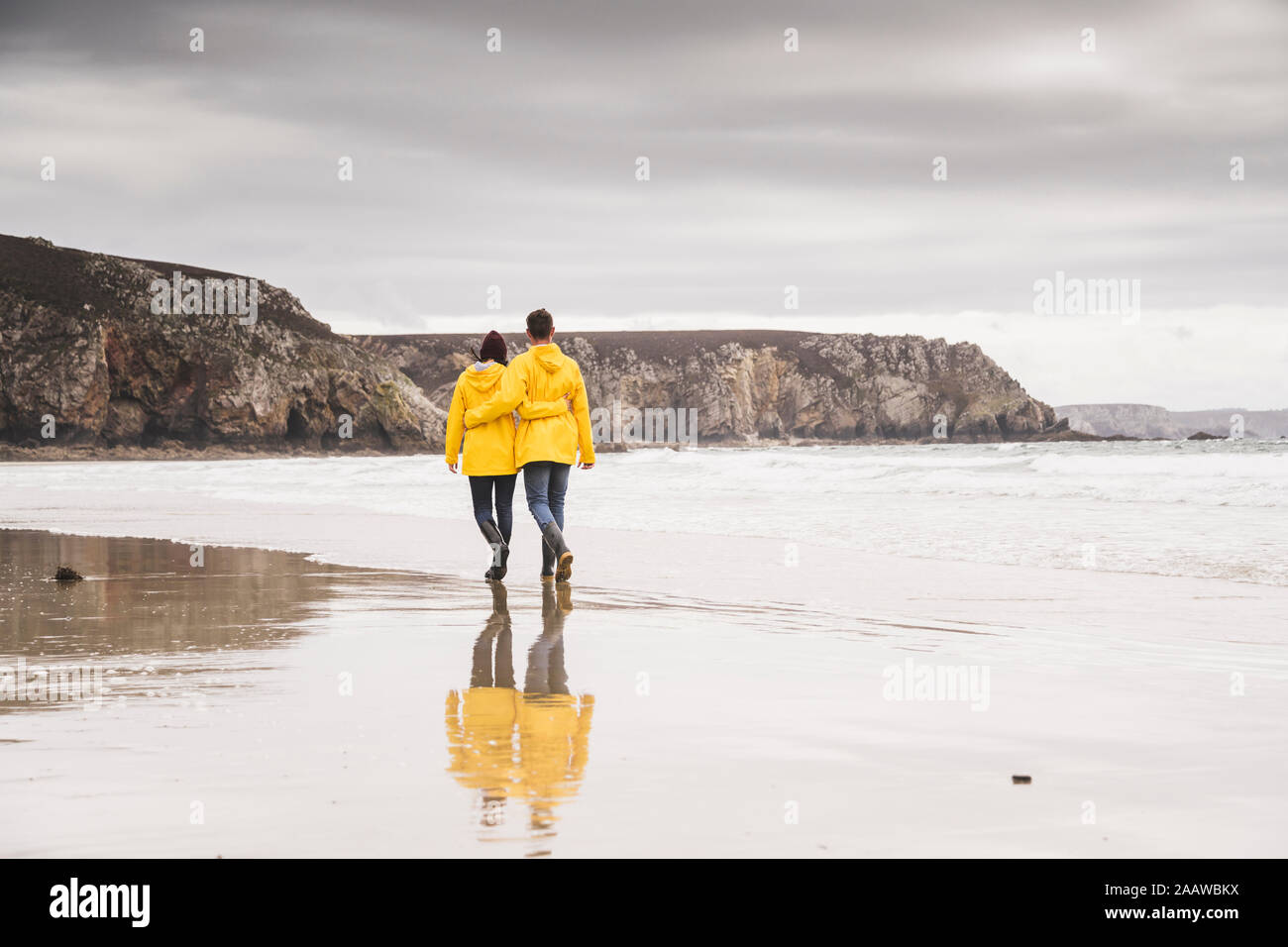 Young woman wearing yellow rain jackets and walking along the beach, Bretagne, France Stock Photo