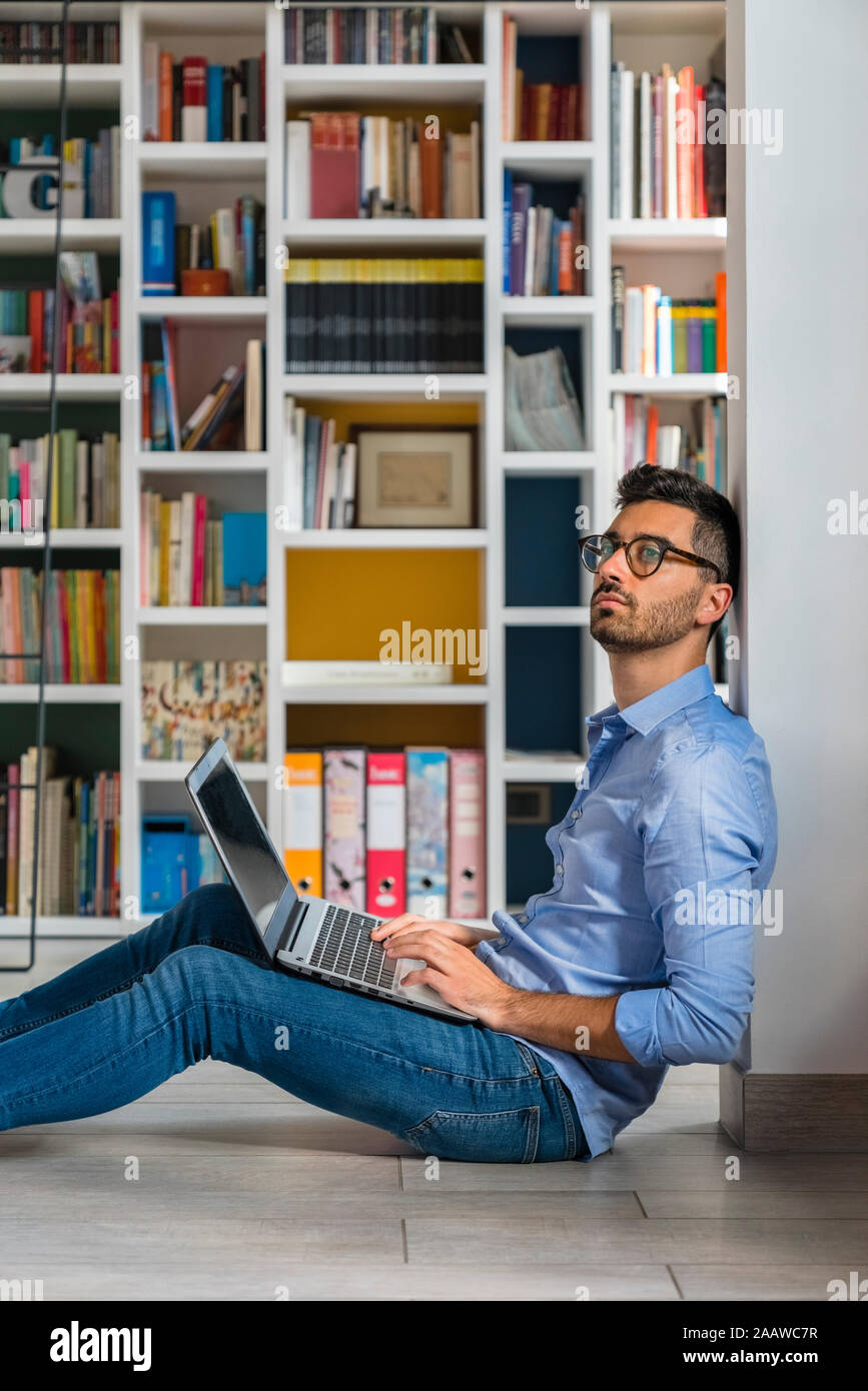Portrait of pensive young man sitting in front of bookshelves on the floor using laptop Stock Photo