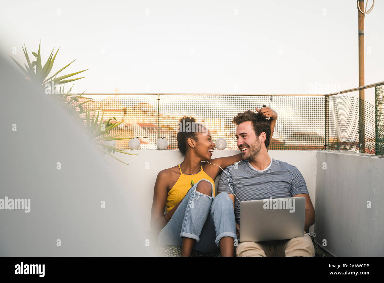 Happy young couple with laptop and earphones sitting on rooftop in the evening Stock Photo