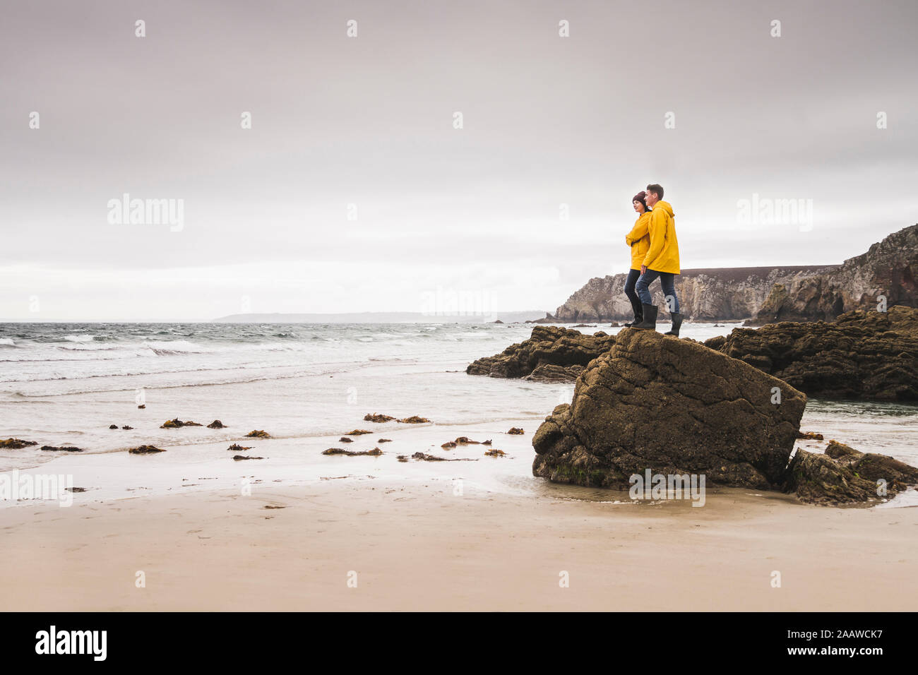 Young woman wearing yellow rain jackets and standing on rock at the beach, Bretagne, France Stock Photo