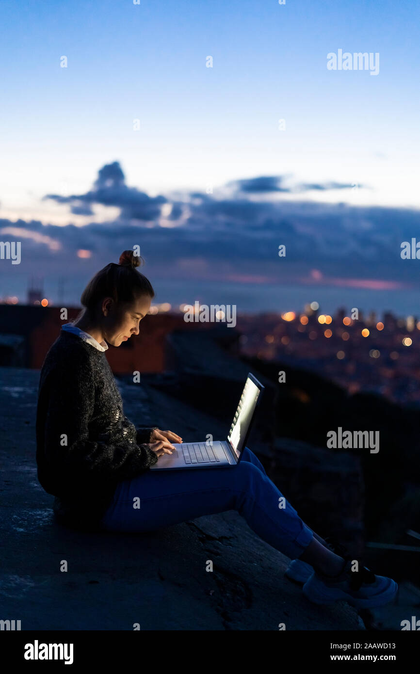 Young woman using laptop at dawn above the city, Barcelona, Spain Stock Photo